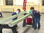 Two children in Cub Scout uniforms blowing toy boats along a race track made of gutters