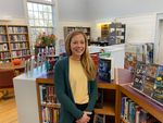 Katie Ball posing for a picture in the library, wearing a cardigan and smiling, with bookshelves, flowers, and a window in the background