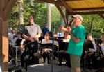 a man n a green shirt and baseball cap speaks into a microphone, next to a band and its director dressed in a black shirt and bow tie