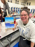 A woman seated at a table showing a painting of an ocean and beach